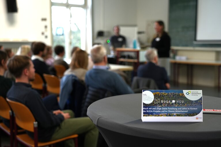 Schild mit Aufruf zur Beteiligung bei BUGA HGU und im Hintergrund die Teilnehmenden im Hörsaal von hinten