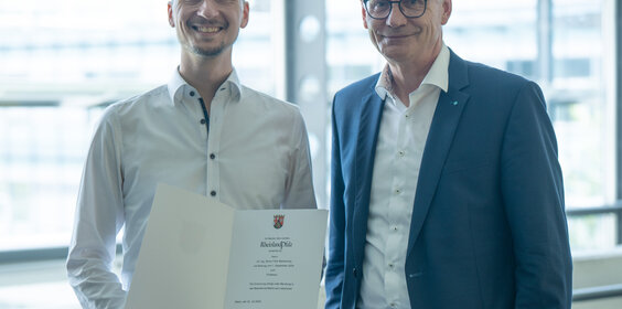 Zwei Herren stehen beieinander. Der Linke von Beiden hält eine Urkunde in der Hand.