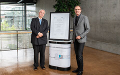 Vertrag zwischen dem DLR Projektträger und der Hochschule Koblenz steht im Rahmen auf einem Tisch. Links daneben steht Prof. Dr. Kristian Bosselmann-Cyran und rechts daneben Klaus Uckel, Gschäftsleiter DLR-Projektträger