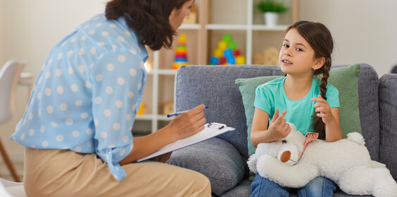 Happy little girl telling story to child psychologist during therapy session in cozy modern office