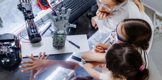 Drei Mädchen und eine Frau sitzen an einem Experimentiertisch. Darauf stehen Roboterhände.