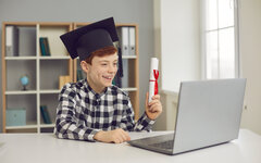 Graduate boy sitting at desk in graduate hat and holding scroll of diploma in front of laptop.