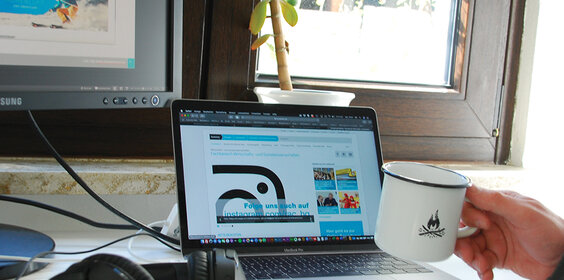 Schreibtisch mit Bildschirm, Laptop und Kopfhörern. Im Vordergrund eine Hand mit einem Kaffeebecher. So sehen Home Office Arbeitsplätze für Studierende aus
