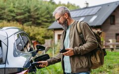 EIn Mann steht mit Handy und Ladekabel an seinem Elektroauto neben einem Haus.