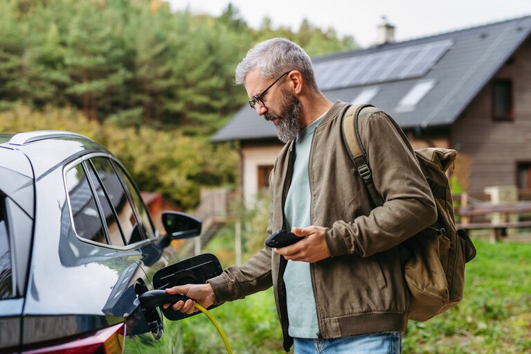 EIn Mann steht mit Handy und Ladekabel an seinem Elektroauto neben einem Haus.